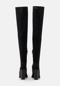 Botas negras por encima de la rodilla con un acabado textil elegante, con un eje ligeramente estructurado y tacones altos en bloque. Disponen de cremalleras laterales para el cierre.