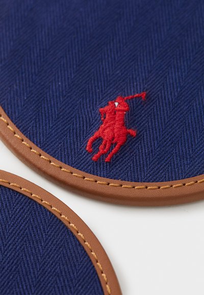 Runde underlag med et marineblått herringbone-mønster, kanter i brunt lær, og en fremtredende rød brodert logo av en rytter.