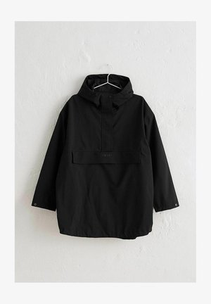 Veste de pluie noire à capuche avec poche avant à rabat, fermetures à boutons-pression au col et aux poignets, suspendue à un crochet mural blanc.