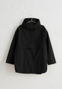 Černá kapucová nepromokavá bunda s předním kapsou s klopou, zapínáním na druky na límci a manžetách, zavěšená na bílém nástěnném háčku.