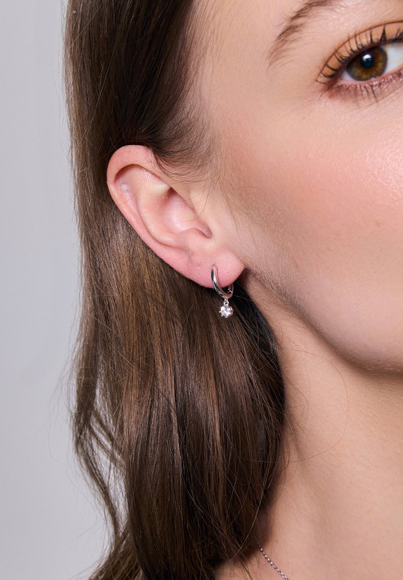 Srebrny kolczyk w kształcie koła z małym kamieniem szlachetnym zawieszonym na dole. Kolczyk ma gładkie, wypolerowane wykończenie i prosty design.