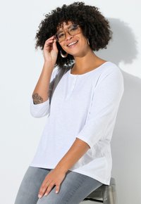 T-shirt blanc à manches longues avec un col rond et une patte de boutonnage à trois boutons. Texture douce, coupe décontractée, associée à un jean gris clair.