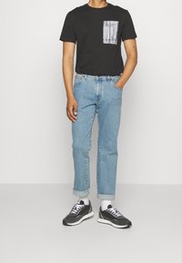 Svart t-shirt med kort ärm och ett mönstrat ficka, ljusblå jeans uppvikta vid fållarna, och svarta sportskor med vita detaljer.