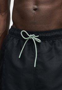 Moški trup s temno kožo v črnih športnih hlačah, zavezanih z svetlo zelenim vrvico, na enobarvni podlagi.