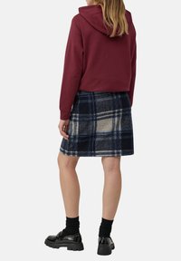 Sudadera recortada en color burdeos con capucha y cordón, combinada con una falda de lana a cuadros en azul marino, crema y gris. Medias negras hasta la rodilla y zapatos brillantes.