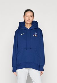 Námornícky modrá mikina s kapucňou a predným vreckom, s vysokým zlatým logom Nike a vyšitým dizajnom na hrudi. Mäkká textúra látky.