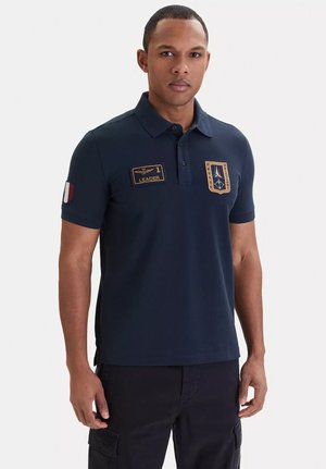 Uomo con polo blu navy con patch "1 Leader" ed emblemi, in piedi davanti a uno sfondo bianco semplice, rivolto in avanti.
