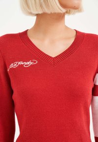 Rot gestrickter Pullover mit V-Ausschnitt, aus gerippter Textur und mit weißem "Ed Hardy"-Logo auf der linken Brust. Lange Ärmel mit kontrastierenden Streifen.