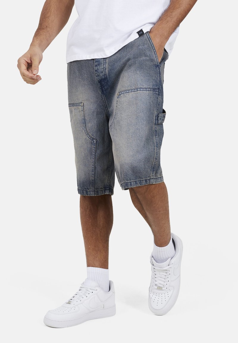 Uomo che indossa pantaloncini di jeans blu sbiadito fino al ginocchio, scarpe da ginnastica bianche e una camicia bianca, con una mano in tasca mentre cammina.