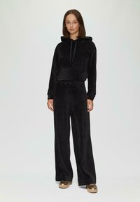 Zwarte velours trainingspak met een hoodie en een voorzak, aangevuld met een elastische tailleband, brede broeken en metallic plateau schoenen.
