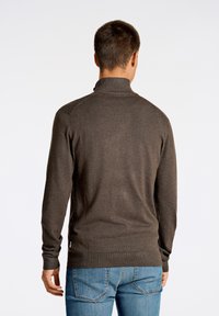 Maglione marrone lavorato a maglia con colletto alto, maniche lunghe e orlo a coste. Indossato con jeans blu, che mostrano una vestibilità aderente e un tessuto texturizzato.