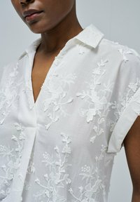 Blusa blanca bordada con patrones florales, mangas cortas y cuello, que muestra un bordado texturizado en una tela ligera.