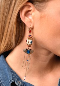 Boucles d'oreilles en ton or avec un cabochon rouge, une pierre précieuse bleue, des détails de perles complexes et des chaînes pendantes avec un design ailé à la base.