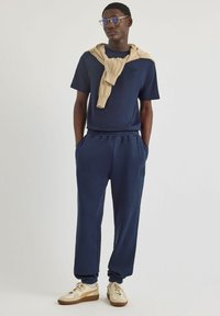 Granatowa bawełniana koszulka z krótkim rękawem z haftowanym logo, w parze ze wzorzystymi spodniami dresowymi oraz beżowym swetrem narzuconym na ramiona.