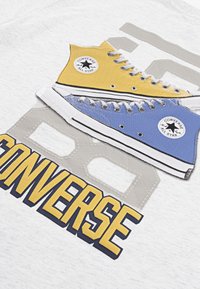 Vit t-shirt med en grafisk design av två sneakers: gula hög-top och blå låg-top Converse, med fet "CONVERSE" text nedanför.