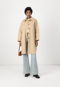 Trench coat beige con colletto, cinque bottoni e tasche laterali. Abbinato a jeans a zampa d'elefante azzurri chiari e scarpe nere. Gli accessori includono una piccola borsa marrone.