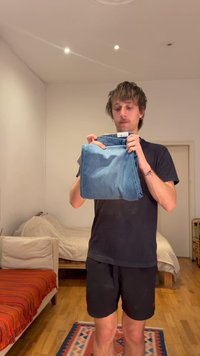 Noor mees voltimas siniseid teksapükse magamistoas, kus on puidust põrand, voodi, diivan punase pleediga ja mustriline vaip.