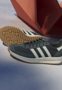 Baskets athlétiques vert foncé et blanc avec semelles en gomme, placées sur un sol clair avec une structure géométrique orange en arrière-plan.