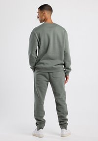 Donkergroene sweatshirt en bijpassende joggers, gemaakt van zacht materiaal, met ribgebreide manchetten en een achterzakdetail, gedragen met witte sneakers.