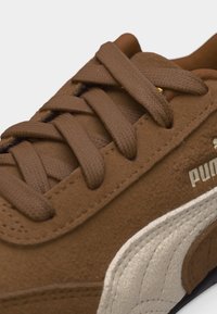 Κοντινή λήψη καφέ σουέτ αθλητικού παπουτσιού με κορδόνια ίδιου χρώματος, μπεζ υφασμένο πλαϊνό ρίγες και μερικό λογότυπο «Puma» στην εξωτερική πλευρά.