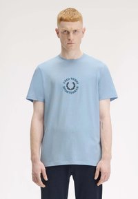 Helles blaues Baumwoll-T-Shirt mit kurzen Ärmeln, das ein schwarzes rundes Logo mit der Aufschrift "FRED PERRY SPORTSWEAR" in der Mitte trägt.
