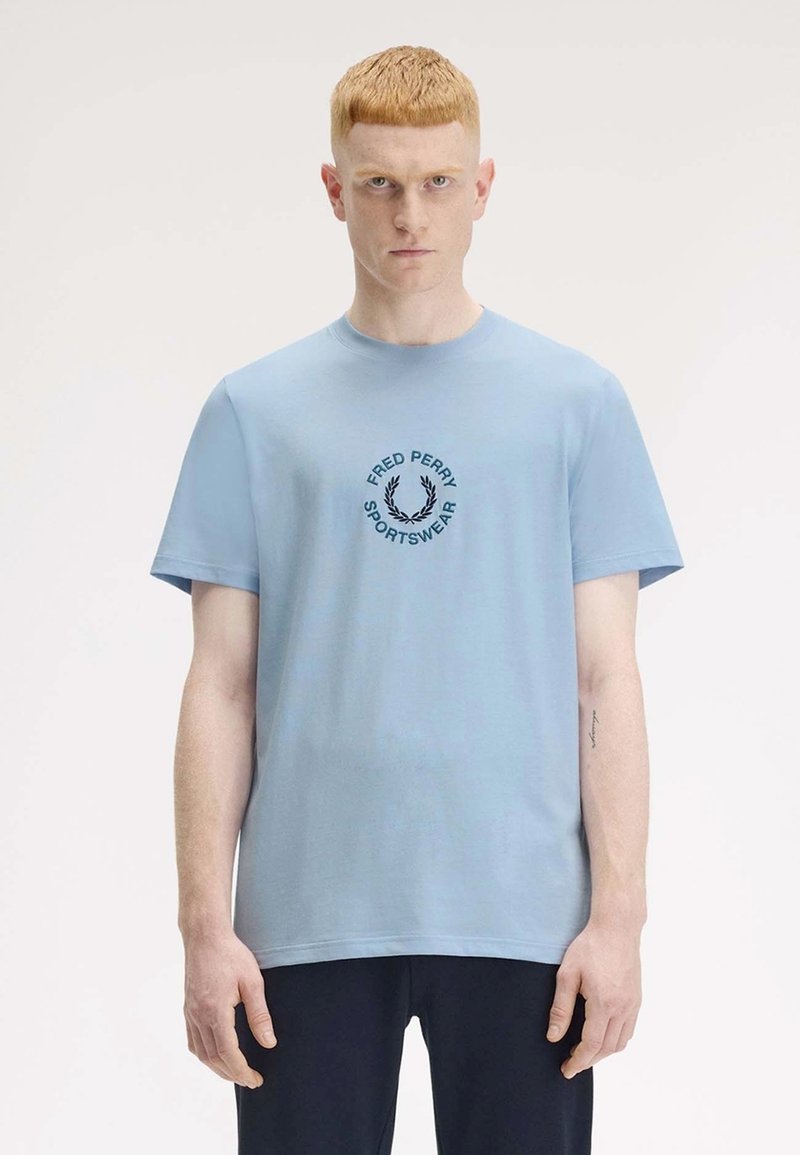 Helles blaues Baumwoll-T-Shirt mit kurzen Ärmeln, das ein schwarzes rundes Logo mit der Aufschrift "FRED PERRY SPORTSWEAR" in der Mitte trägt.