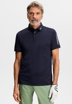 Uomo anziano in piedi che indossa una polo blu navy con logo sul colletto e sulla manica, pantaloni verde chiaro e un guanto da golf bianco sulla mano destra.