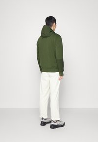 Grön bomullshuvtröja med huva, ribbade muddar och nederkant. Bärs med vita byxor och grå vandringsskor med svarta sulor och röda detaljer.