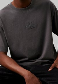 Muž v tmavě šedém tričku Calvin Klein Jeans a černých kalhotách, sedí s jednou rukou opřenou o stehno.