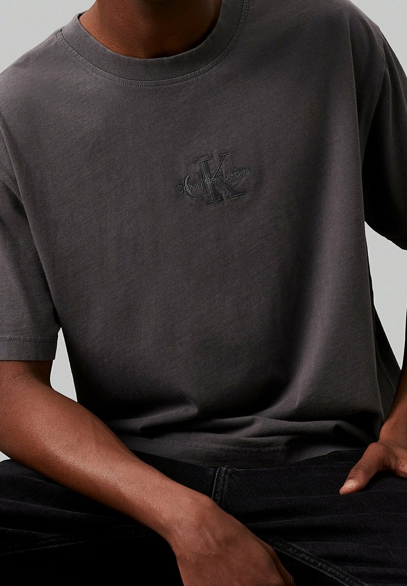 Muž v tmavě šedém tričku Calvin Klein Jeans a černých kalhotách, sedí s jednou rukou opřenou o stehno.