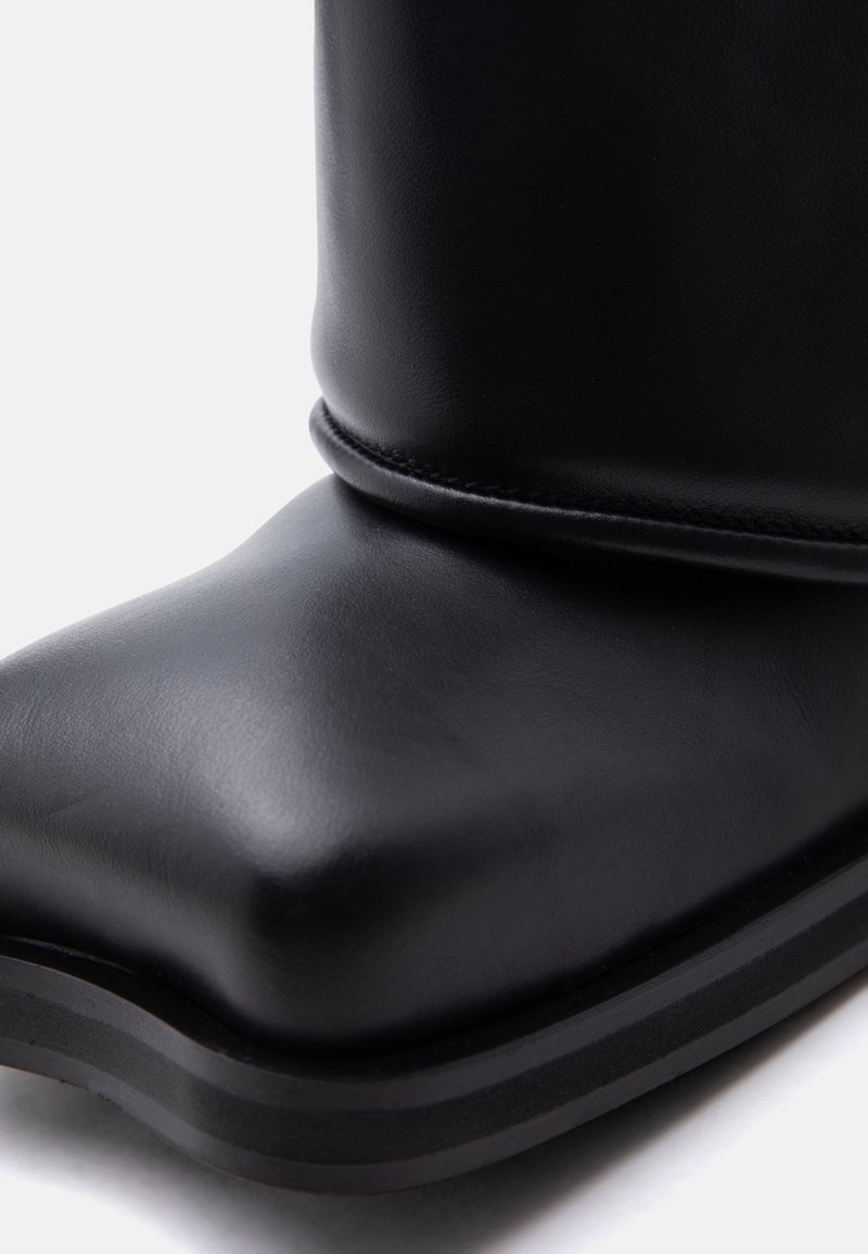 Close-up of a black leather boot with a square toe, folded upper edge, and a thick layered sole.