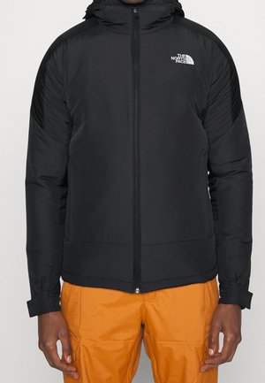Veste matelassée noire zippée avec capuche et logo The North Face sur la poitrine, portée avec un pantalon orange.