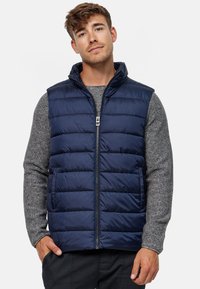 Marineblauer gepolsterter Weste mit hohem Kragen, Frontreißverschluss und horizontalem Steppmuster, getragen über einem grauen strukturierten Pullover.