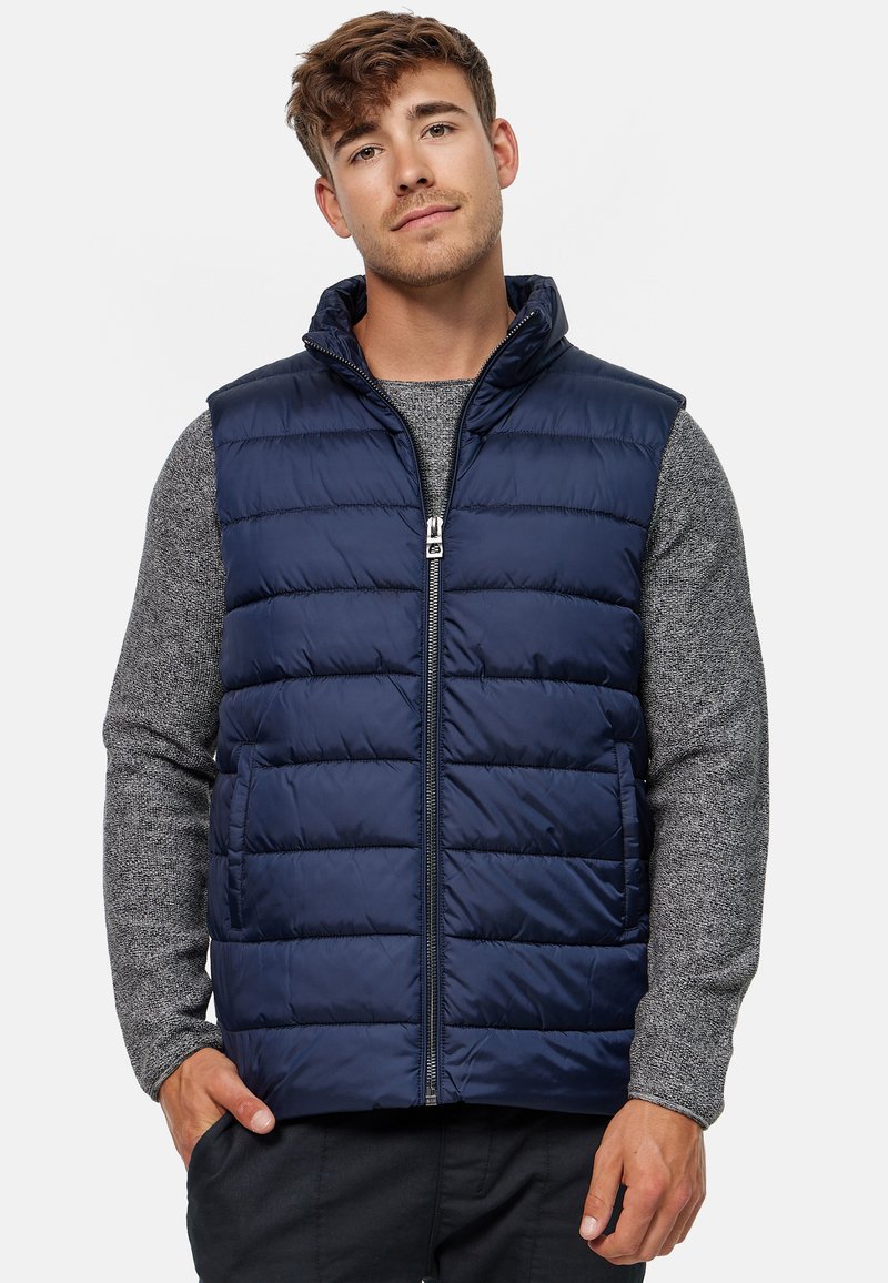 Gilet imbottito blu navy con colletto alto, zip frontale e design trapuntato orizzontale, indossato sopra un maglione grigio a trama.