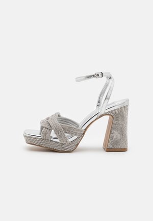 Srebrne sandały na wysokim obcasie z wiązanym paskiem z przodu i masywnym obcasem zdobionym. Posiadają metaliczne wykończenie i zapięcie na kostkę.