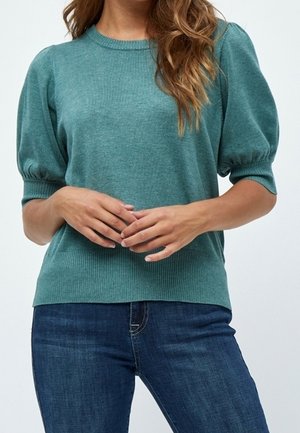 Frau trägt einen türkisfarbenen Strickpullover mit Puffärmeln und dunkelblaue Jeans, Hände locker auf der Taille verschränkt vor einfarbigem Hintergrund.