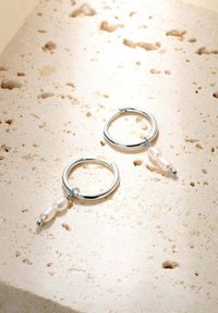 Des boucles d'oreilles en argent présentent deux perles roses et un pendentif en perle argentée, exposées sur une surface en pierre beige texturée.