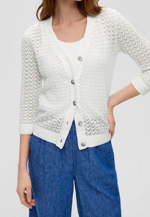 Vrouw draagt wit kantgebreid vest met knopen en driekwart mouwen over witte top en blauwe spijkerbroek, hand op heup.