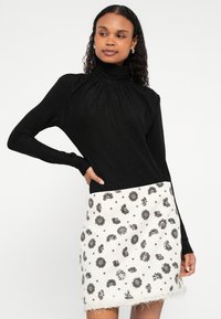 Crna majica dugih rukava s imitacijom rolke i naboranim izrezom uparena s bijelom mini suknjom ukrašenom srebrnim cvjetnim uzorcima i resicama na rubu.