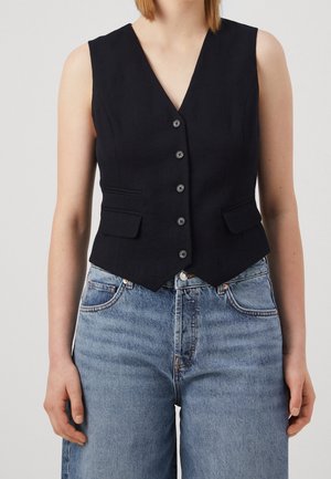 Nainen, joka pukeutuu hihattomaan mustaan napitettuun liiviin, jossa on kaksi etutaskua, yhdistettynä vaaleansinisiiin korkeavyötäröisiin farkkuihin.