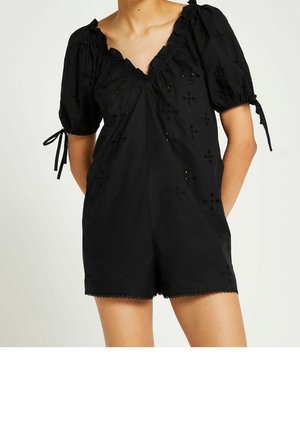 Kobieta ubrana w czarny kombinezon z bufkami, haftem w oczka, falbaniastym dekoltem i związanymi końcami rękawów, stojąca z rękami za plecami.