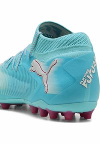 Botas de fútbol azul claro con una parte superior texturizada, cuello de punto y acentos rosas en la suela. Logo exhibido en el costado.