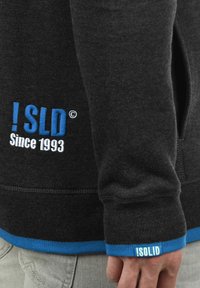 Sweat-shirt noir avec des accents bleus, arborant un logo brodé "! SLD Since 1993" à l'avant et un ourlet côtelé, fabriqué en tissu doux.