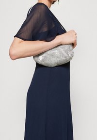 Silverfärgad paljettclutch hålls vid midjan, har en rundad form och mjuk textur. Bärs med en flödande marinblå klänning.