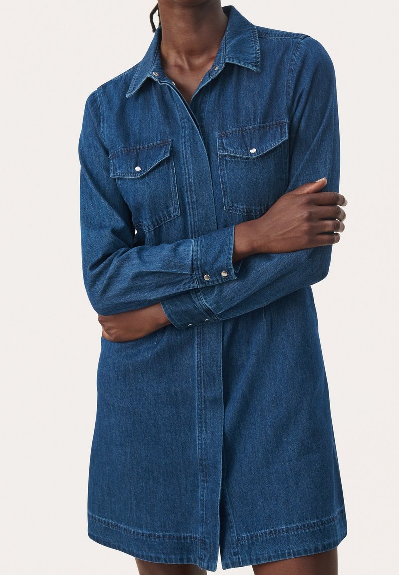 Personne portant une robe en denim bleu à manches longues avec des poches poitrine boutonnées, debout les bras croisés devant un fond clair uni.