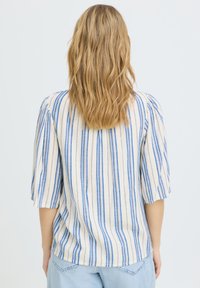 Femme aux cheveux blonds ondulés portant un chemisier blanc à rayures verticales bleues et un jean bleu clair, de dos sur un fond uni.