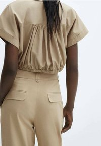 Blusa color tan con maniche corte e dettagli arricciati sul retro, abbinata a pantaloni alti color tan con due tasche posteriori.