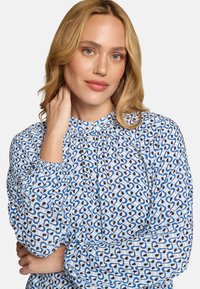 Blouse bleu et blanc à motif de cœurs, avec des manches froncées et une fermeture boutonnée sur le devant. Tissu léger avec une texture lisse.