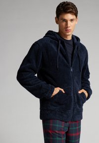 Námořnická modrá mikina na zip vyrobená z měkkého, nadýchaného materiálu; má kapuci, přední kapsy a žebrované manžety. Nositá přes kostkované kalhoty.