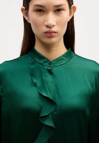 Grüne Satinbluse mit Mandarin-Kragen und Rüschendetail an der Blende. Glatte Textur mit glänzendem Finish. Einziger schwarzer Knopfverschluss.